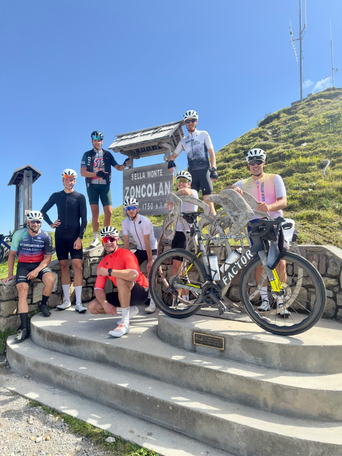 Nine riders in helmets and kit posing with their bikes at the Monte Zoncolan summit sign (1,750 m) on a sunny day.